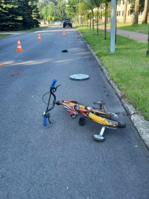 Na jezdni stoją pachołki odgradzające miejsce zdarzenia drogowego, w którym znajduje się rower dziecięcy, kołpak i czapka, w oddali pojazdy.